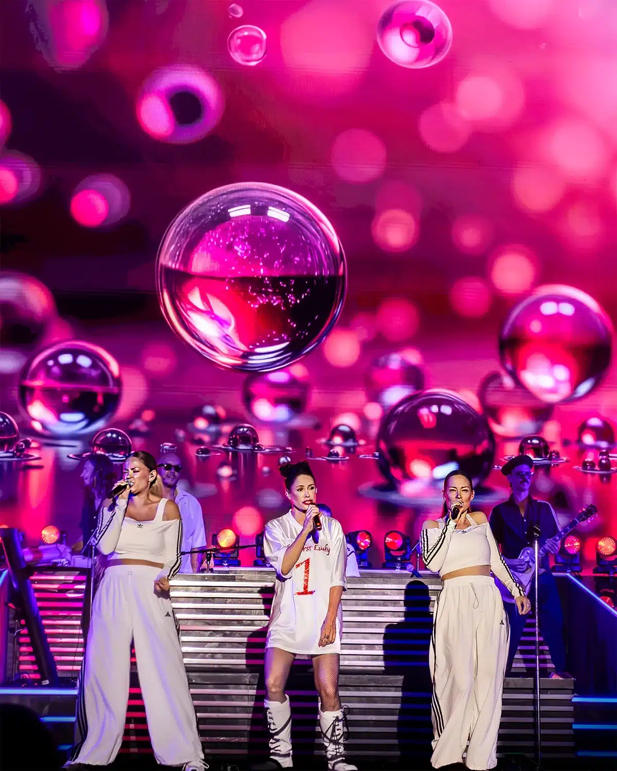 AQUA Lene Nystrøm på scenen med korsangere, iført hvid trøje med teksten The First Lady, foran lyserød boble-grafik.