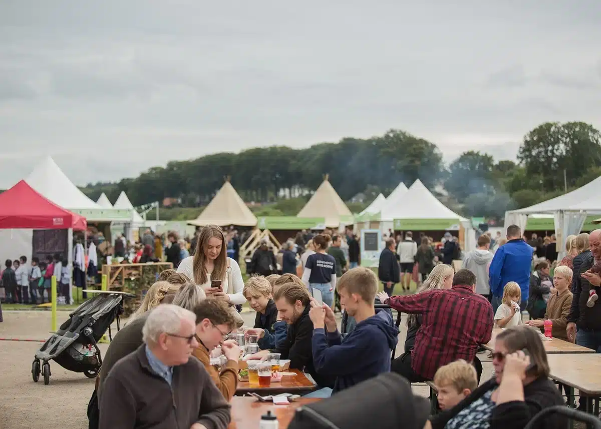 Gæster sidder ved langborde og nyder mad og drikke på Food Festival Gæster sidder ved langborde og nyder mad og drikke på Food Festival