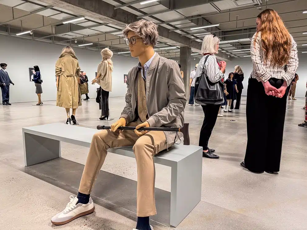 Ung mannequin med gangstok sidder på bænk blandt besøgende på udstillingen The Populace på ARoS