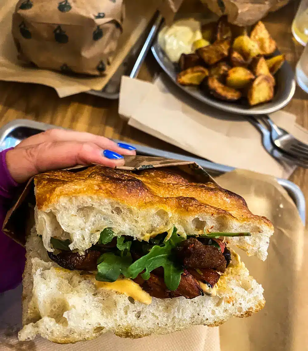 BBQ mushroom focacciasandwich med rucola og mayo holdt i hånden på Plant Food med ovnstegte kartofler i baggrunden
