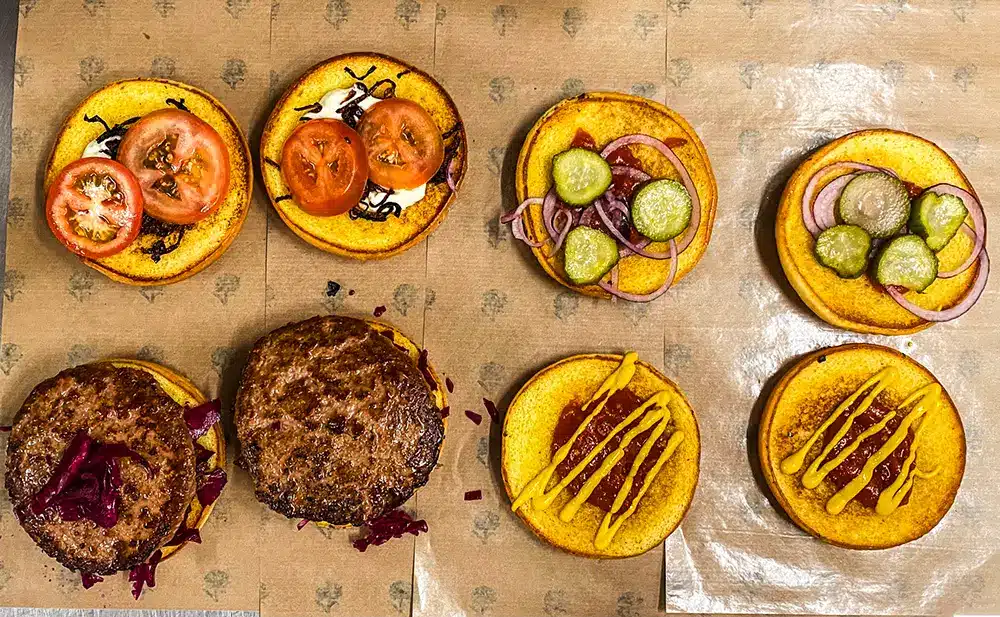 Plantebaserede burgere bliver samlet i Plant Foods køkken med briocheboller, tomat, stegte løg, syltede rødløg, agurk, ketchup og sennep